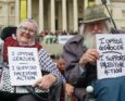 Due persone anziane sorridenti sulla sedia a rotelle mostrano il cartello in inglese con la scritta “Mi oppongo al genocidio. Sostengo Palestine Action”