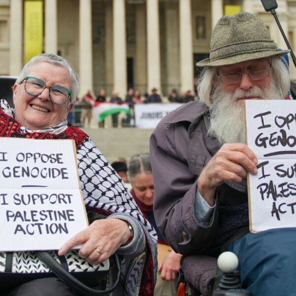 Due persone anziane sorridenti sulla sedia a rotelle mostrano il cartello in inglese con la scritta “Mi oppongo al genocidio. Sostengo Palestine Action”