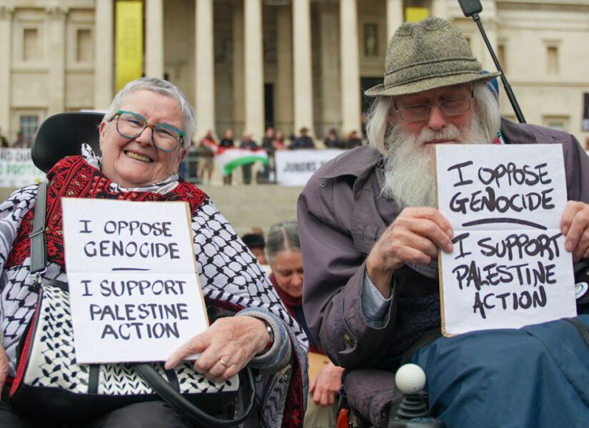 Due persone anziane sorridenti sulla sedia a rotelle mostrano il cartello in inglese con la scritta “Mi oppongo al genocidio. Sostengo Palestine Action”