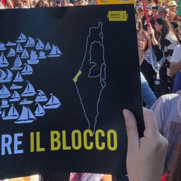 Un cartello "Rompere il blocco" con delle piccole barche che formano un pesce grande verso la Strscia di Gaza. Dietro la folla alla manifestazione di Roma del 4 ottobre 2025