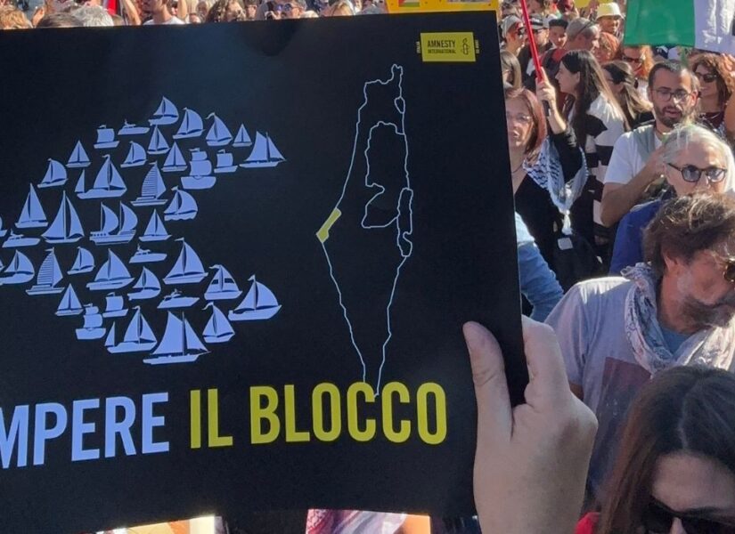 Un cartello "Rompere il blocco" con delle piccole barche che formano un pesce grande verso la Strscia di Gaza. Dietro la folla alla manifestazione di Roma del 4 ottobre 2025