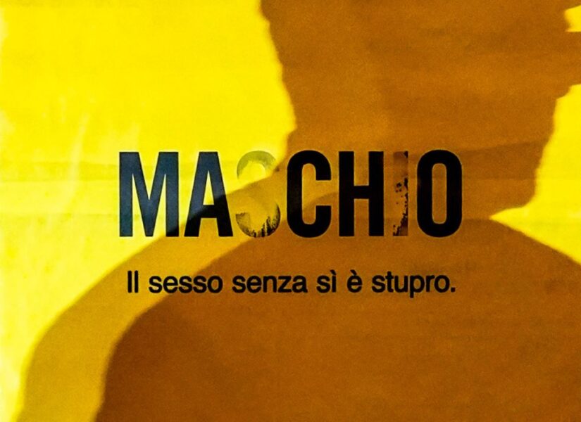 L'ombra di un uomo si staglia su un manifesto giallo con la scritta nera "Maschio" in cui le lettere che compongono il sì spariscono trasformando la parola in "macho"